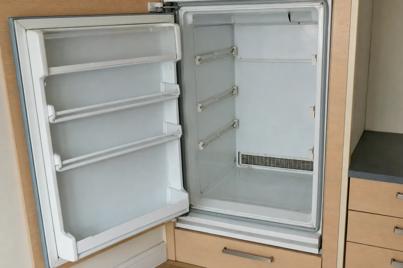 A built-in undercounter refrigerator set into pale wood cabinetry with its door open, showing an empty white interior and the vent grille low on the back wall