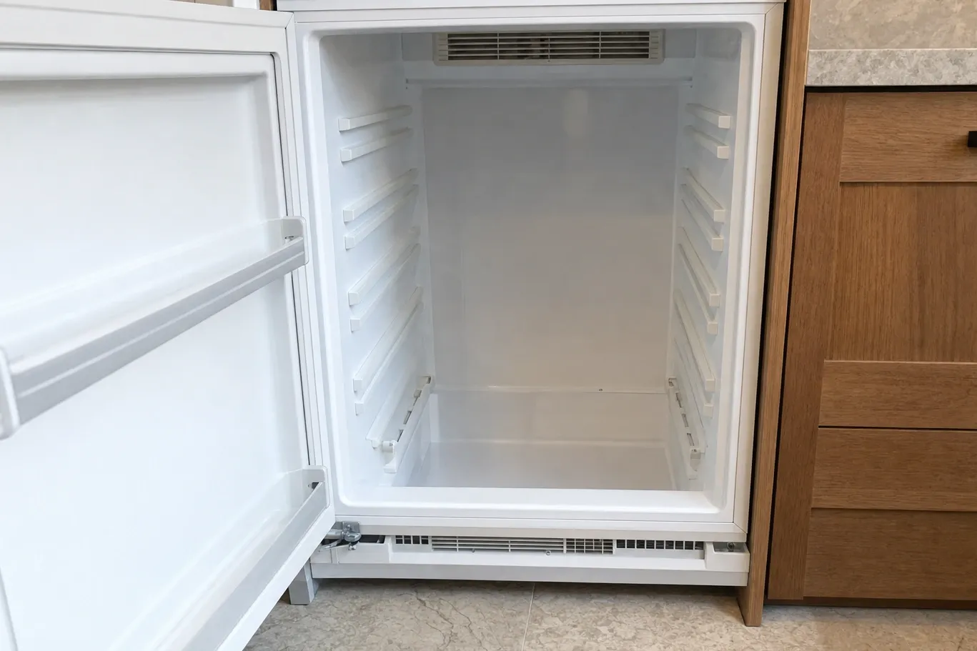 An undercounter refrigerator with its door open on an empty white interior and the base vent grille removed and standing on the floor in front of it