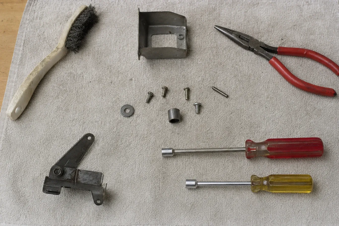 A door hinge assembly, a folded sheet-metal shield, a wire brush, long-nose pliers, two nut drivers and loose screws laid out on a canvas drop cloth