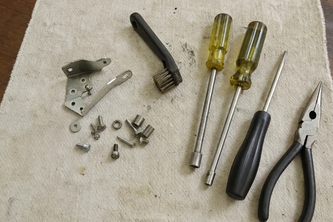 A stainless hinge with its latch arm beside a wire brush, two nut drivers, a scratch awl and combination pliers on a canvas cloth, with screws and standoffs grouped alongside