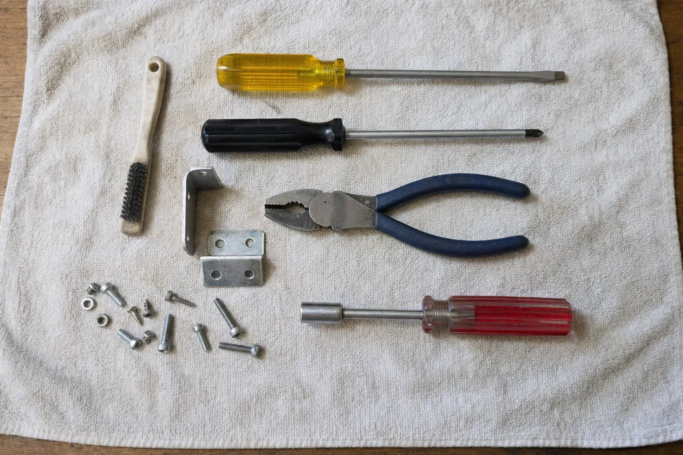 An angle bracket and a flat mounting plate laid out on a towel with two screwdrivers, a nut driver, combination pliers, a wire brush and a scatter of machine screws and nuts