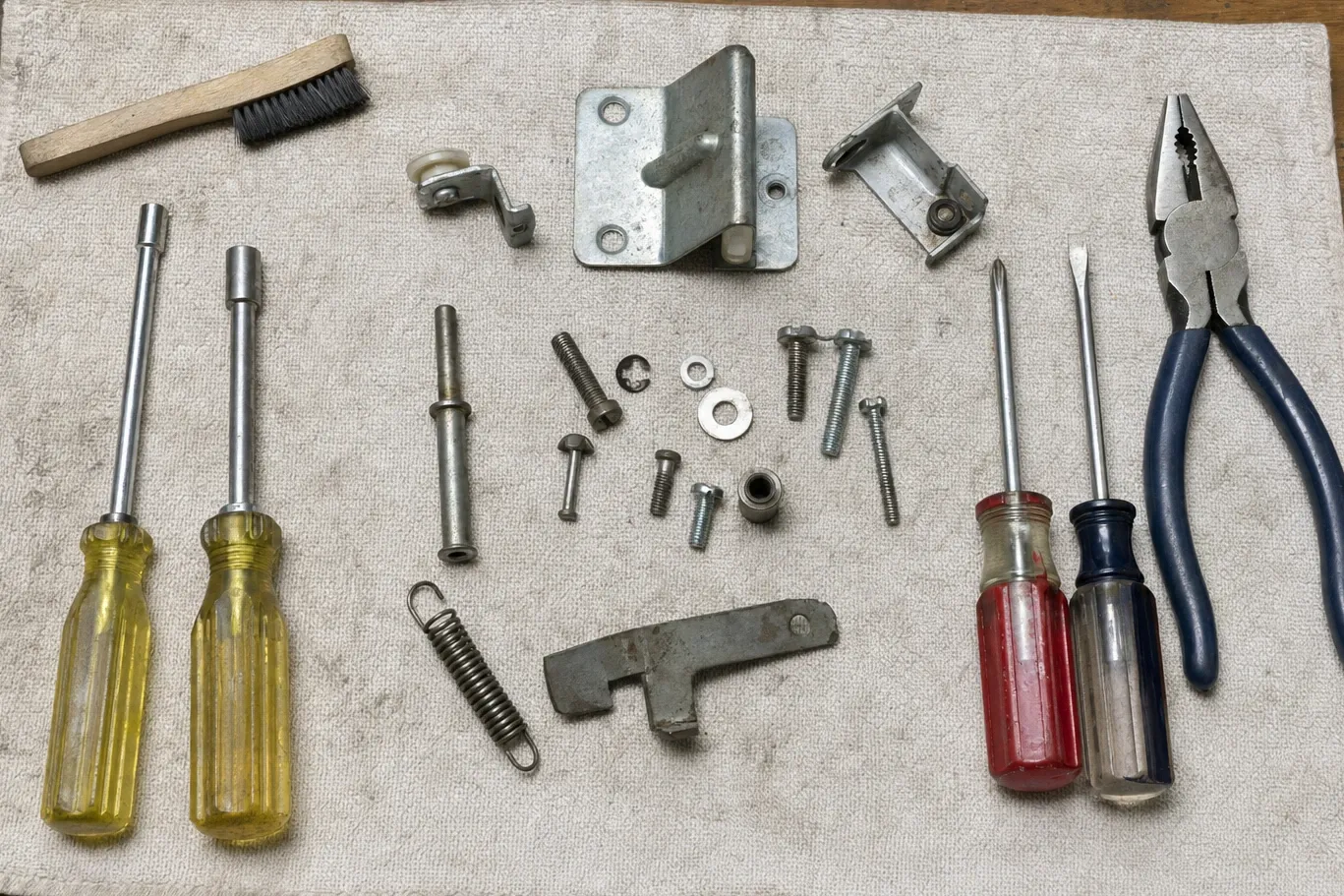 A heavy hinge plate with its pivot pin, a latch striker, a tension spring and a latch arm laid out on canvas with nut drivers, screwdrivers, pliers and a bench brush